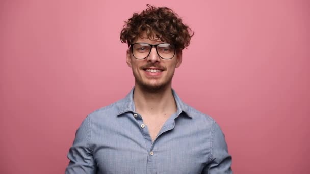 mladý ležérní muž nosí brýle stojící a dívá se na kameru šťastný pak stoupající ruce nahoru vzrušený na růžové pozadí studia - Záběry, video