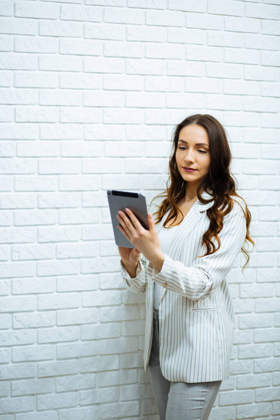 Улыбающаяся деловая женщина с темными волосами в белом брючном костюме держит планшет в руках работает полноразмерные расчеты возле белой стены
. - Фото, изображение