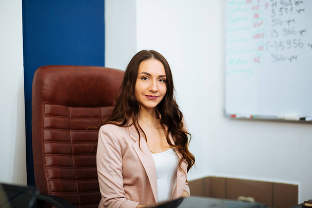 smiling beautiful business brown-haired lady with long hair sitting at the desk on an office chair looking ahead career growth. - Foto, imagen