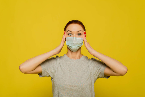 A woman clings to her head and experiences fear, shock from the spread of the disease. Red-haired beautiful girl in a protective mask on a yellow background screams from a coronavirus. The concept of protection and prevention. - Photo, Image