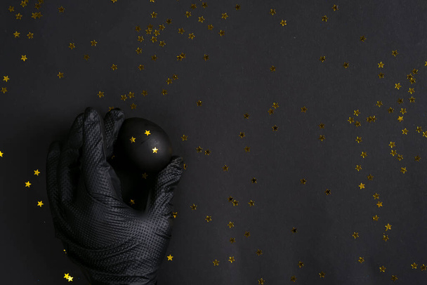 Womans hand in a dark glove holds easter handmade black painted egg on the same color background with golden confetti decoration. - Photo, Image