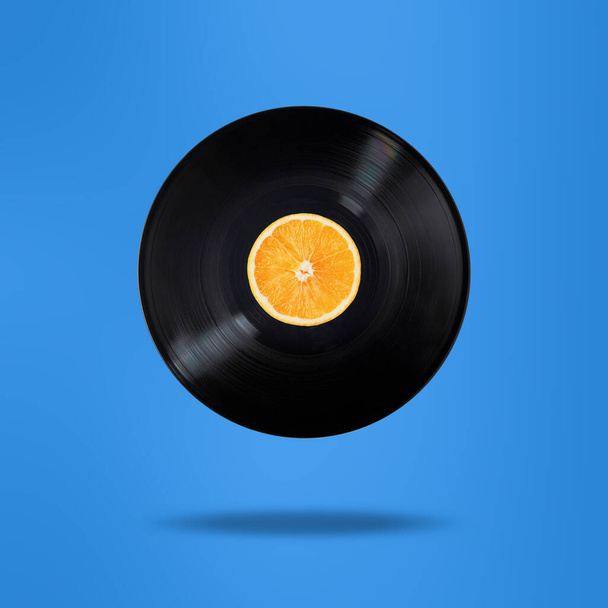 Vinyl ρεκόρ με πορτοκαλί φρούτα στο κέντρο. Πετάω στον αέρα. Απομονωμένο σε μπλε φόντο. Σουρεαλιστική έννοια ανύψωσης. - Φωτογραφία, εικόνα