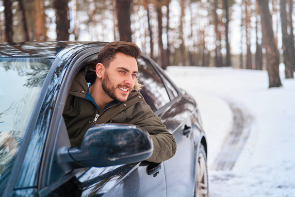 Young adult attractive Caucasian man sits at the wheel of his car sunny winter day. Wintertime road trip. Happy smiling hipster guy sitting in car and looking window. Portrait Positive driver  - Photo, image