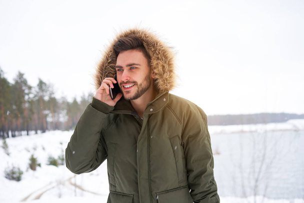 Handsome smiling brutal bearded young caucasian man standing outdoors fur hood in winter season forest Talking on a cell phone Attractive stylish european guy walking snowy christmas landscape - Foto, Imagen