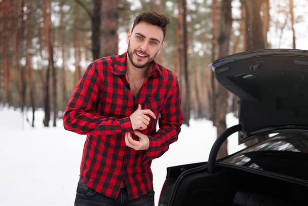 Young adult handsome man standing winter forest near his car. Attractive caucasian guy waiting for help when car is broken. Winter landscape road trip.  - Foto, Bild