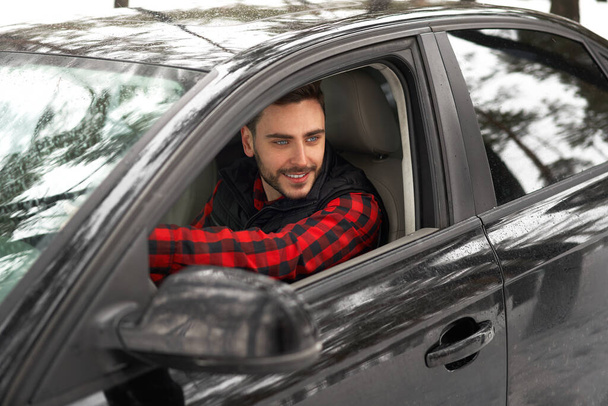 Young adult attractive Caucasian man sits at the wheel of his car sunny winter day. Wintertime road trip. Happy smiling hipster guy sitting in car and looking window. Portrait Positive driver  - Фото, зображення