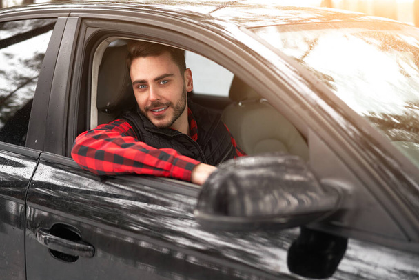 Young adult attractive Caucasian man sits at the wheel of his car sunny winter day. Wintertime road trip. Happy smiling hipster guy sitting in car and looking window. Portrait Positive driver  - Photo, image