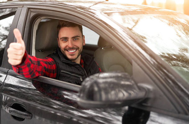 Young adult attractive Caucasian man sits at the wheel of his car sunny winter day shoeing thumbs up gesture. Wintertime road trip. Happy smiling hipster guy sitting in car and looking window.  - Fotó, kép