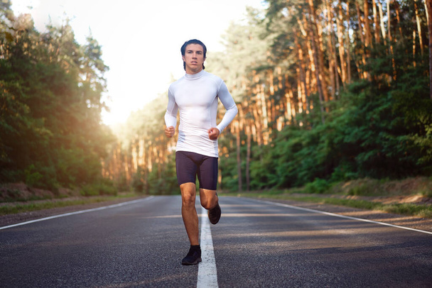 Caucasian middle age man athlete in white t shirt runs sunny summer day on  asphalt road in the pine forest. Men 30s years jogging at the nature. training for marathon run. Healthy lifestyle concept. - Fotoğraf, Görsel