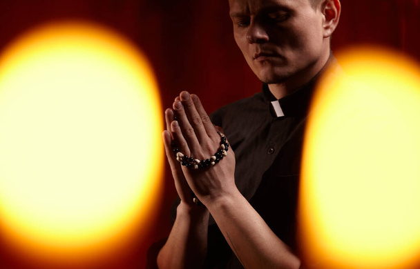 Young adult caucasian catholic priest on dark red background Handsome pastor father pray indoors in dark authentic church - Fotografie, Obrázek