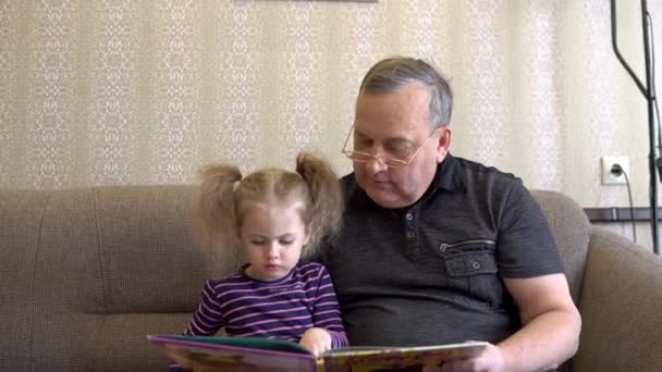 Granddaughter is reading a book with grandfather. The girl frowns at the book and listens carefully to Grandfather. Sitting on the couch together closeup - Filmati, video