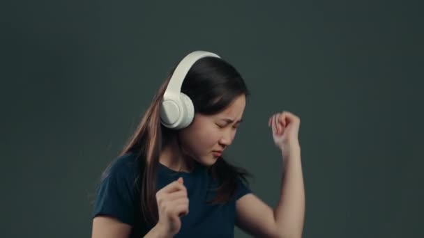 Pretty young asian girl with long hair having fun, smiling, dancing with headphones in studio against grey background. Music, dance, radio concept. - 映像、動画