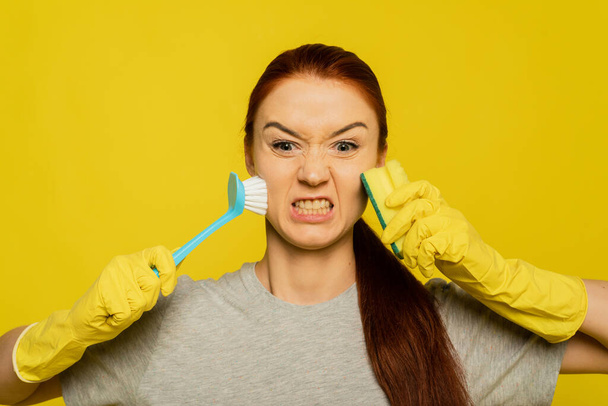 An angry woman with an aggressive grimace holds a pipe and a brush for washing dishes. An excited girl rubs her skin very much, doing make-up removal and skin cleansing. Evil emotion on the face with a grin. - Photo, Image