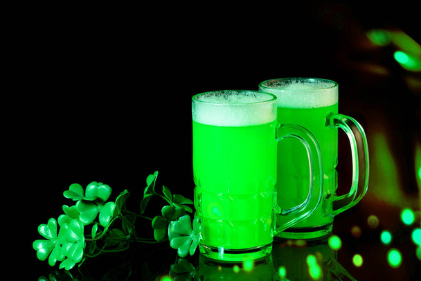 Día de San Patricio tradicional fiesta irlandesa. Tazas de cerveza verde con hojas de trébol sobre el fondo negro reflectante. Luces, buen bokeh, diseño de arte
.  - Foto, imagen