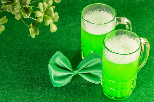 Duas canecas de vidro de cerveja verde, folhas de trevo e gravata borboleta como símbolos do Dia de São Patrício no fundo verde. Festival irlandês tradicional
.  - Foto, Imagem