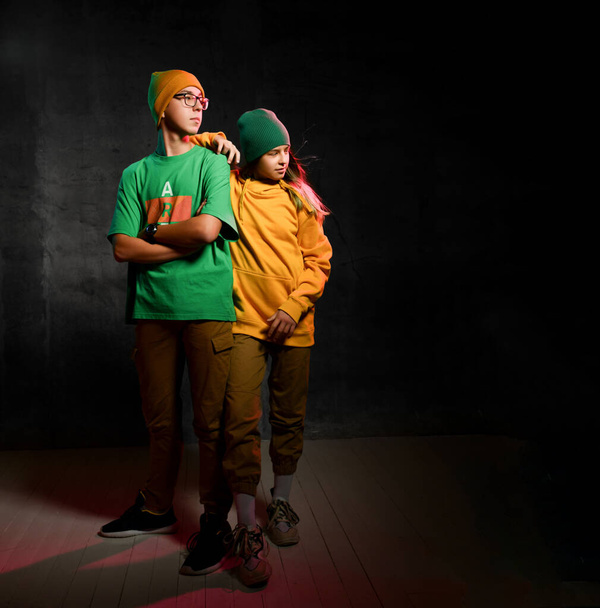 Young teens boy and girl in green and yellow comfortable casual clothing and hats standing and looking aside over dark background - Valokuva, kuva