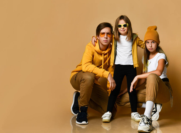 Young teens and small girl in comfortable casual clothing and sunglasses sitting and looking at camera over yellow background - Foto, afbeelding