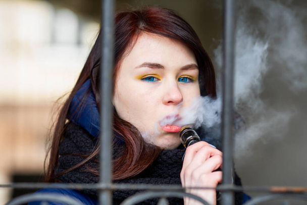 Vape teenager. Young pretty white caucasian brunette girl with blue eyes and in a blue jacket smoking an electronic cigarette near old metal gate on the street in the spring. Deadly bad habit. - Foto, imagen