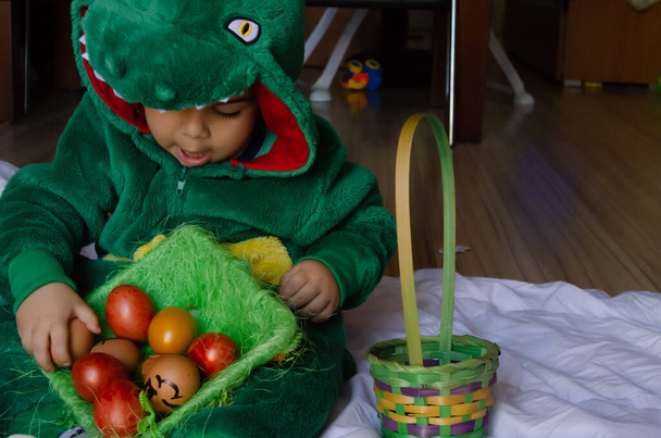 Милый малыш в зеленом костюме динозавра играет с красочными пасхальными яйцами
.. - Фото, изображение