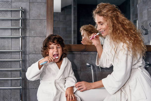 Ελκυστική χαμογελαστή νεαρή μητέρα με ρόμπα καθαρίζει τα δόντια της μαζί με όμορφο μικρό γιο κοντά νιπτήρα με καθρέφτη. Οικογενειακή έννοια. - Φωτογραφία, εικόνα