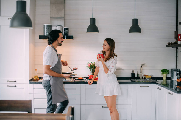 International Women's Day. Young couple in the morning in the kitchen at home. A man cooks a woman a festive breakfast on March 8 in a beautiful stylish kitchen. - Foto, afbeelding