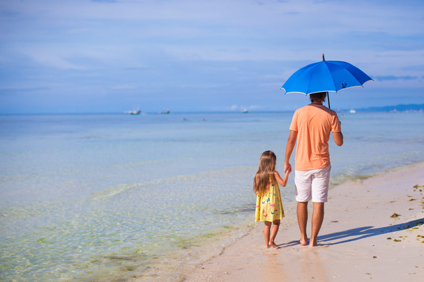 Nuori isä ja hänen suloinen pieni tyttärensä piilossa auringolta sinisen sateenvarjon alla valkoisena aurinkoisena päivänä.
 - Valokuva, kuva