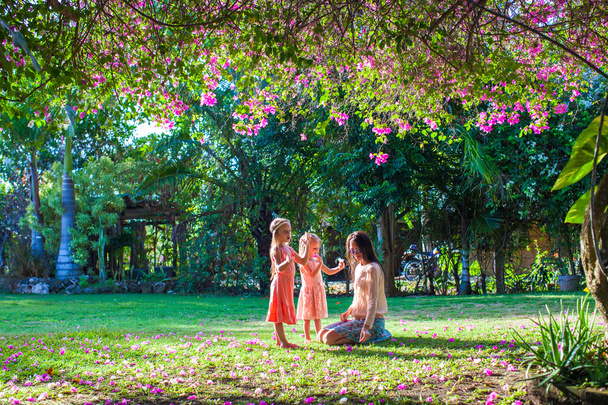 Молода мати зі своїми маленькими доньками сидить у пишному саду і насолоджується літнім відпочинком
 - Фото, зображення