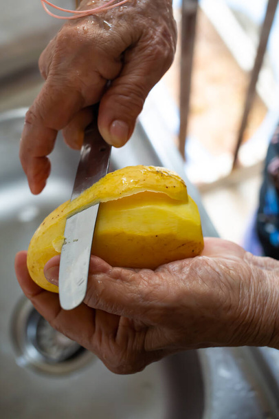 Die rechte Hand der Seniorin mit einem Messer, das süße gelbe Mango im Spülbecken schält, Nahaufnahme, selektiver Fokus, traditionelle thailändische Früchte - Foto, Bild