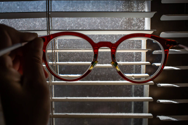 Mão de mulher segurando óculos míopes ou míopes com persianas de poeira dentro de uma janela, poeira e mosquito sujo janela de tela de arame, Light & Shadow tiro, tiro de close-up, foco seletivo
 - Foto, Imagem