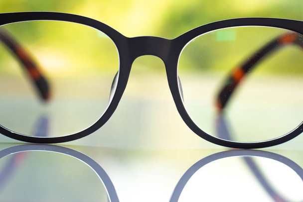 óculos de visão curta ou míope preto na mesa de acrílico branco, fundo de jardim verde Bokeh, Close up & Macro tiro, foco seletivo, Reflexão, conceito óptico
 - Foto, Imagem