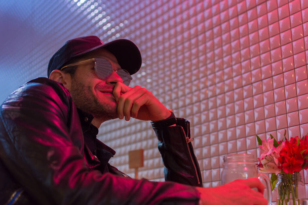 Young latin man portrait with cap, leather jacket and sun glasses with his hand over his mouth in a place with neon lights. Shallow depth of field - Photo, Image