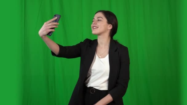 Bella ragazza bruna in un abito da lavoro scatta foto di se stessa su una fotocamera smartphone. Lo sfondo è uno schermo verde per la digitazione
. - Filmati, video