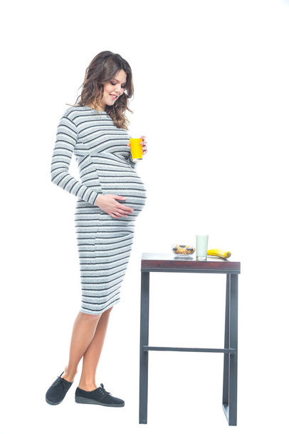A young pregnant woman sits at a table on which lies a banana, there is a plate with cereal, a glass with kefir and juice. The concept of nutrition during pregnancy. Isolated on a white background. - Фото, изображение
