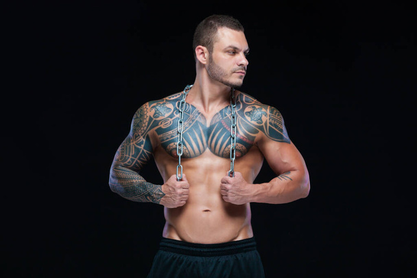 Tattooed strong muscular athletic man holding metal chain over his neck on black background - Foto, Bild