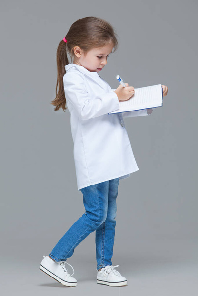 Adorable child girl uniformed as doctor is writing medical record isolated on grey background - Fotografie, Obrázek