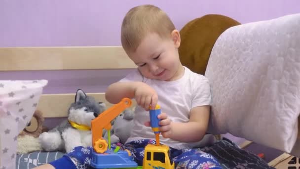 Bájos baba ül az ágyon, és csavarja és forgatja a csavarokat egy játékautóban.. - Felvétel, videó
