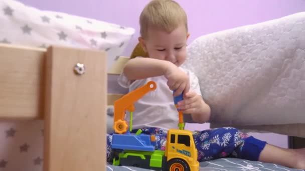 魅力的な赤ちゃんはベッドの上に座って紆余曲折し、おもちゃの車のボルトを回します. - 映像、動画