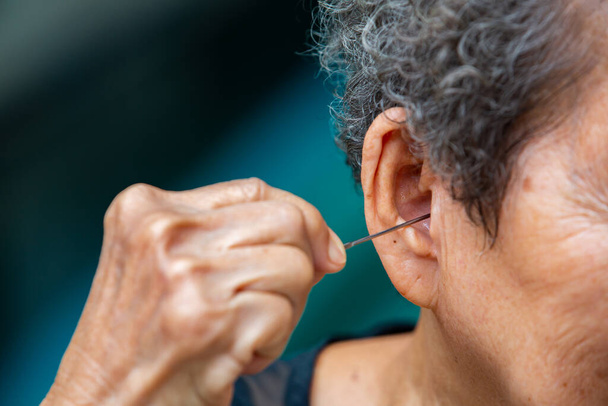Senior mulher colocando earpick em seu ouvido direito, cabelos encaracolados cinzentos, fundo piscina, Close up & Macro tiro, foco seletivo, parte do corpo asiático, conceito de cuidados de saúde, Sintoma de perda auditiva
 - Foto, Imagem