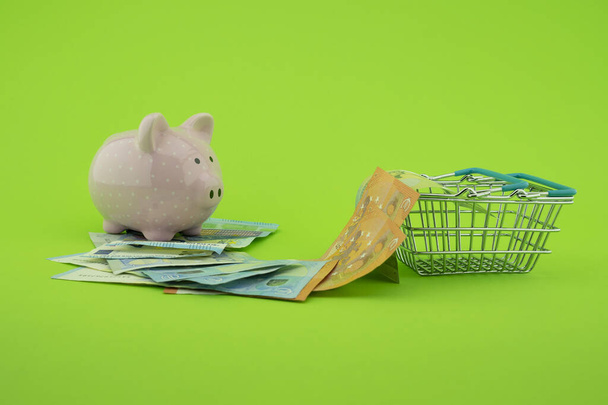 Cost of shopping or spending savings concept with banknotes scattered on a green background with a piggy bank and small wire shopping basket - Photo, Image