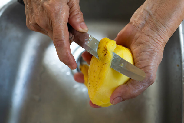 Δεξί χέρι ηλικιωμένης γυναίκας χρησιμοποιώντας μαχαίρι ξεφλούδισμα γλυκό κίτρινο μάνγκο σε φόντο νεροχύτη, Κοντινό πλάνο, επιλεκτική εστίαση, Ταϊλάνδης παραδοσιακά φρούτα - Φωτογραφία, εικόνα