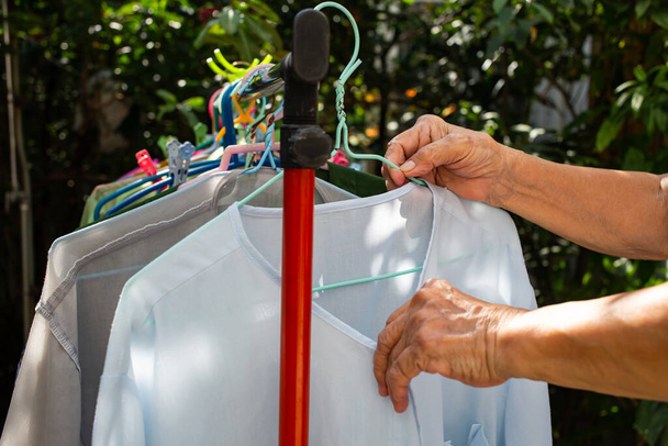 Senior γυναικείο χέρι στεγνό μπλε ρούχα στον ήλιο με πολύχρωμα πλαστικά ρούχα κρέμονται σε σχάρα ρούχα από ανοξείδωτο χάλυβα, Επιλεκτική εστίαση, Πλυντήριο έννοια εργασίας - Φωτογραφία, εικόνα