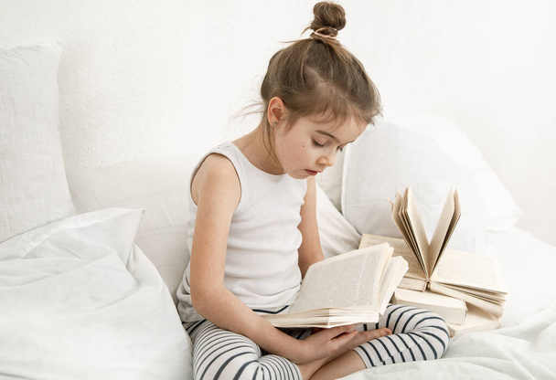 Cute little girl reading a book on the bed in the bedroom. The concept of education and family values . - Foto, Imagem