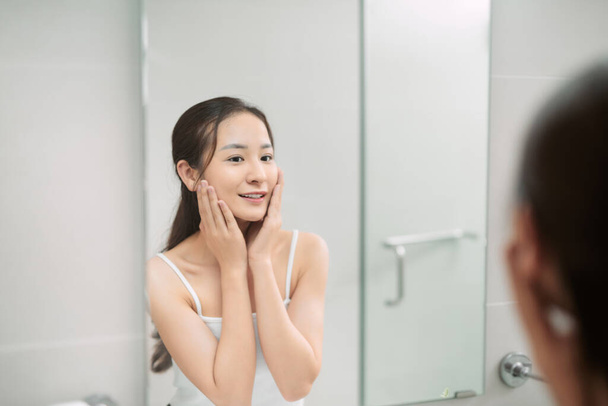 beauty, skin care and people concept - smiling young woman applying cream to face and looking to mirror at home bathroom - Photo, Image