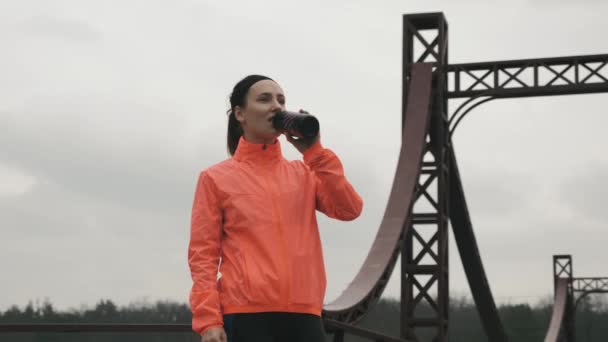 Female athlete is drinking water outdoor in park. Young sportive woman in bright sportswear drinks energy drink from bottle before training. Sports and running concept - Кадри, відео