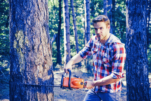 Strong man lumberjack with an axe or chainsaw in a plaid shirt. A handsome young man with a beard carries a tree. - Fotó, kép