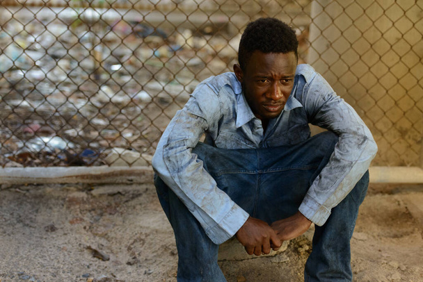 Portrait of young homeless African man against chain link fence in the streets outdoors - Photo, Image