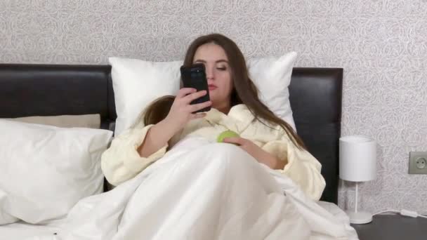Plus size young woman lying in bed, messaging in smartphone, switching tv channels - Imágenes, Vídeo