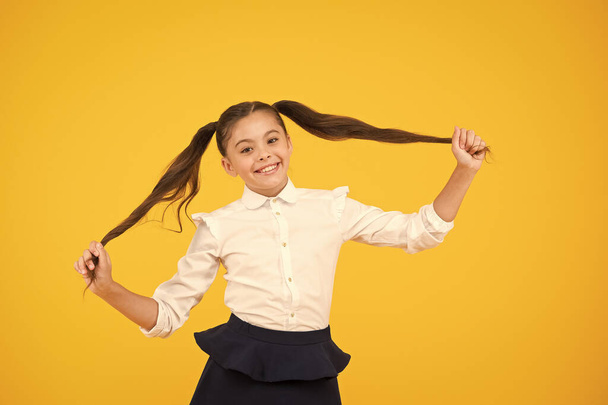 Long and healthy. Cute small schoolgirl holding long hair ponytails on yellow background. Adorable little child in formal wear being proud of long hair. Styling long hair for school - Photo, image