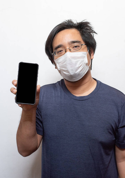 Aziatische jongeman in casual dragen chirurgische medische masker Covid-19 te beschermen met behulp van slimme mobiele telefoon geïsoleerd op witte gezondheidszorg concept.Wuhan coronavirus (Covid-19) uitbraak preventie in de openbare ruimte.  - Foto, afbeelding
