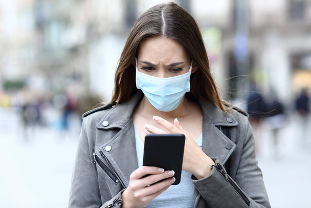 Front view portrait of a scared girl wearing protective mask avoiding contagion reading news on her smart phone on a city street - Fotografie, Obrázek